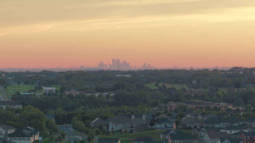 Telephoto aerial of a leafy Minnesota suburb at sunset, with new family homes and winding streets in the foreground and the downtown Minneapolis skyline hazy on the horizon.