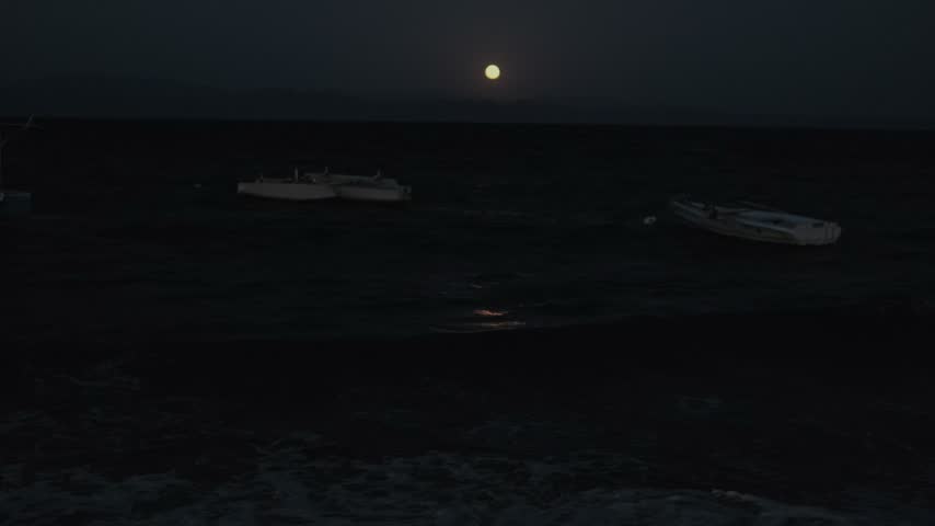 Full moon over rough ocean waves at night with boats and moonlight reflection