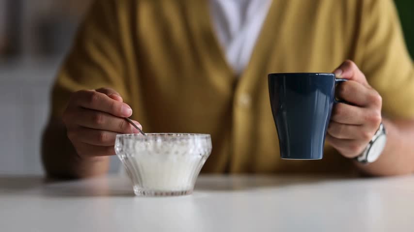 Man enjoying breakfast in a bright kitchen, holding a cup of coffee in one hand and adding a spoonful of sugar. Calm and inviting morning atmosphere.