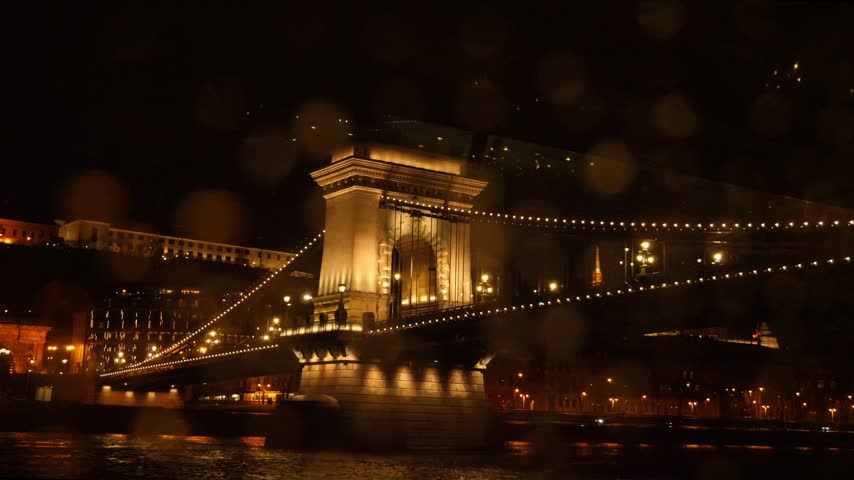 Night view of the illuminated Széchenyi Chain Bridge crossing the Danube in Budapest, Hungary, surrounded by glowing city lights and historic riverside architecture.
