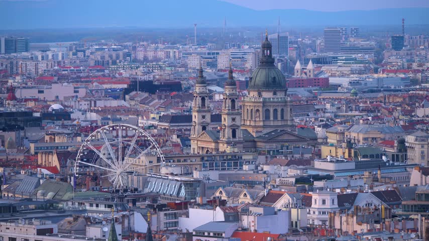 Elevated panoramic view over central Budapest, Hungary, featuring St. Stephen’s Basilica, a Ferris wheel, dense historic buildings and distant hills