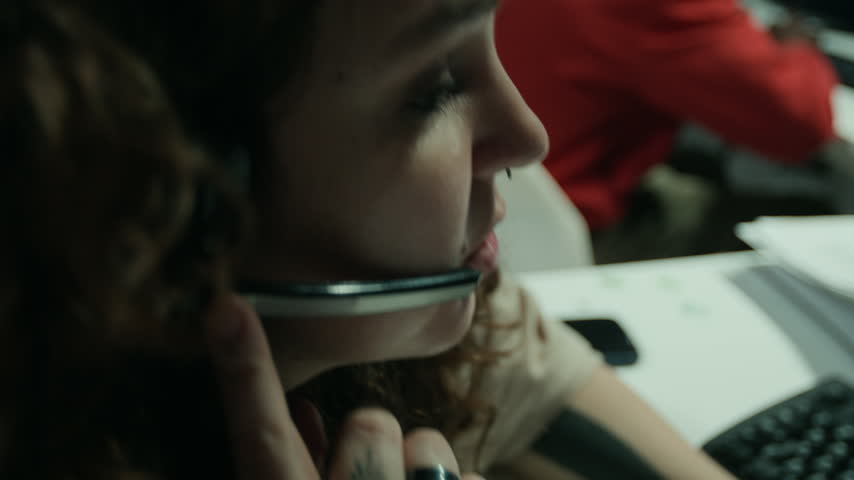 Young Caucasian woman wearing headset, speaking into microphone and taking notes at busy call center desk. Ideal for customer service, tech support or corporate communication projects