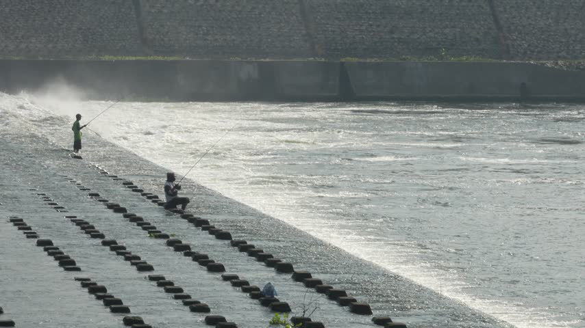 Two fishermen positioned at different levels on concrete steps of a dam structure, engaged in outdoor river fishing.