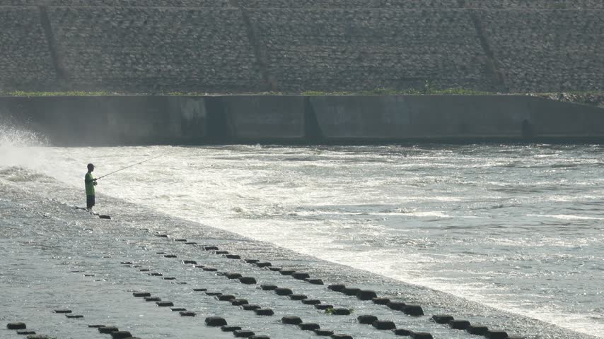 A single fisherman stands holding a rod on concrete blocks near a flowing river dam with water mist under bright daylight.
