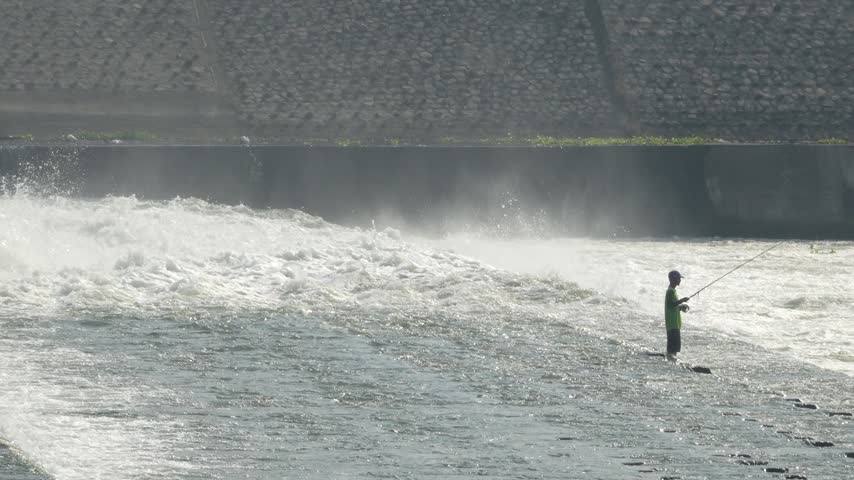 A closer shot of an angler holding a fishing rod near a powerful, rushing, and misty dam waterfall.