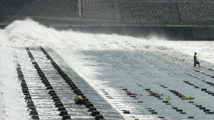 Two distant fishermen positioned on separate tiers of concrete blocks along a flowing river dam during a sunny day.