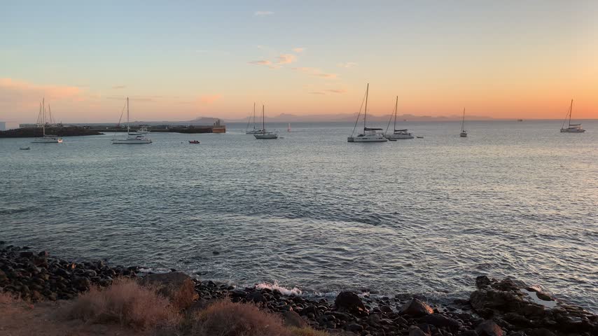 Sunset over the Atlantic Ocean in Playa Blanca, Lanzarote, with sailing boats, volcanic rocky coastline and golden reflections on the water, Canary Islands, Spain.