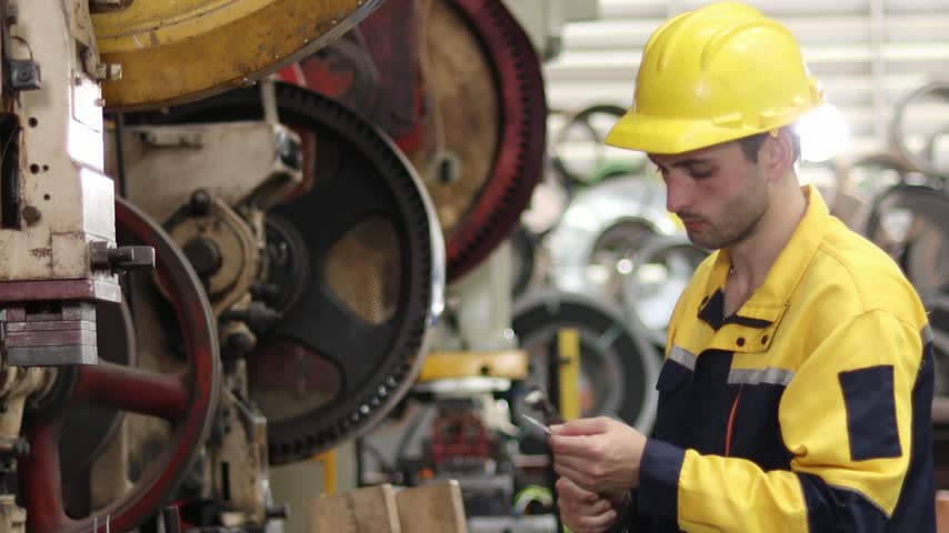 Young worker with safety clothing and helmet inspecting workpiece while working with machine at industrial manufacturing factory. Engineer technician man operating machinery in industrial factory.