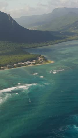 Dramatic mountain peaks rise above turquoise lagoon on Mauritius, sandy peninsula with small resort curves into calm water beneath layered clouds and light coastal mist. Aerial drone vertical shot
