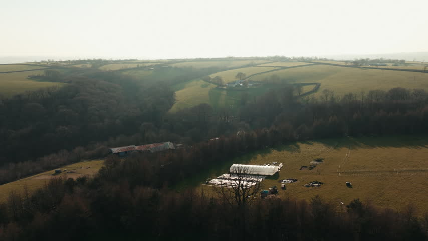 A sweeping aerial shot shows rolling hills and farmland with a long farm building nestled in a misty valley.