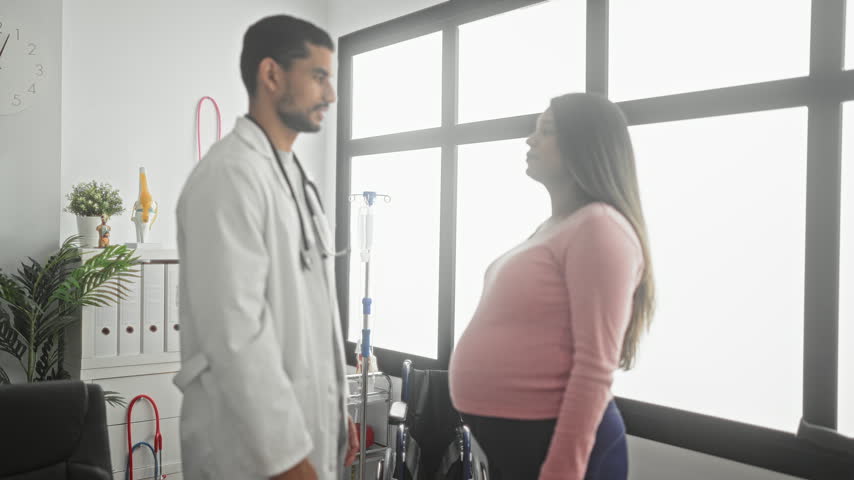Pregnant woman hugging doctor wearing a white coat and stethoscope in a clinic building by a large window and clock; support relief.
