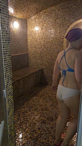 Woman stands in mosaic sauna, smiling at camera. She wears blue bikini top and purple head covering. Warm lighting highlights her in the steam-filled room. Glossy tiles line the walls
