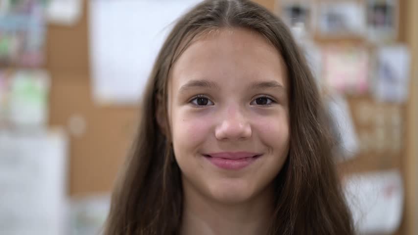 Happy 13-year-old schoolgirl portrait, long brown hair, warm smile, looking at camera in classroom.