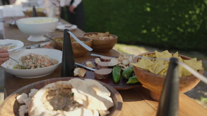 An outdoor buffet on a timber table with tortilla chips, salads, bread and dips in wooden bowls, guarded by battery fly repellent fans.