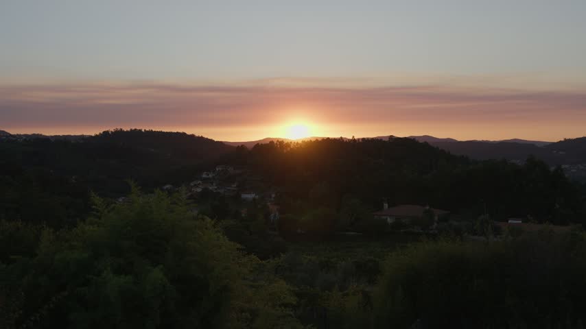 The sun sets behind wooded hills, leaving an orange glow in the sky over a village and dark forest below.