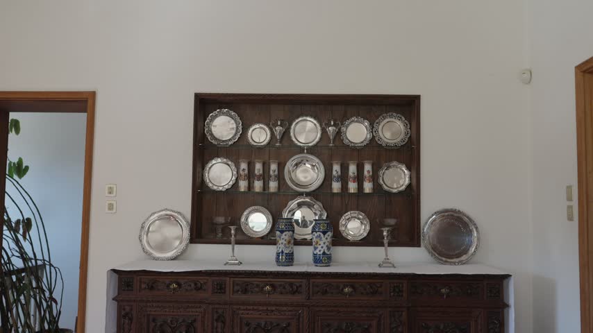 Antique silver platters displayed in a wall niche above a carved wooden sideboard, with blue and white vases in a bright room.