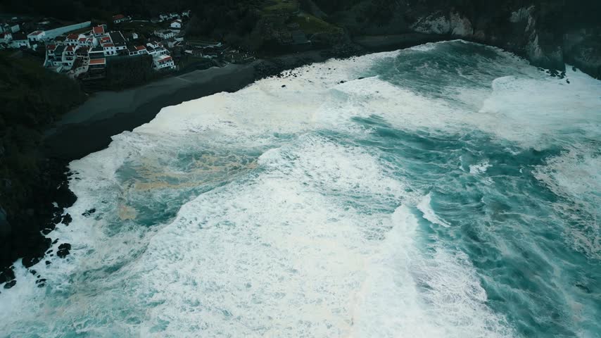 Aerial view of waves rolling onto a rocky shoreline, revealing the rugged coast and a small town perched above the beach. Dynamic movement of water highlights natural beauty.