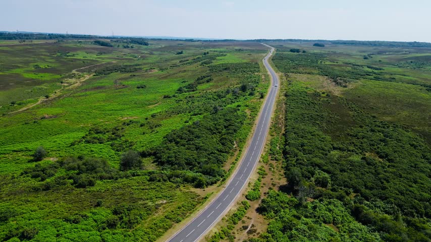 Aerial view of paved road winding through expansive green heathland and woodland with distant rural countryside reaching clear horizon across New Forest Hampshire England