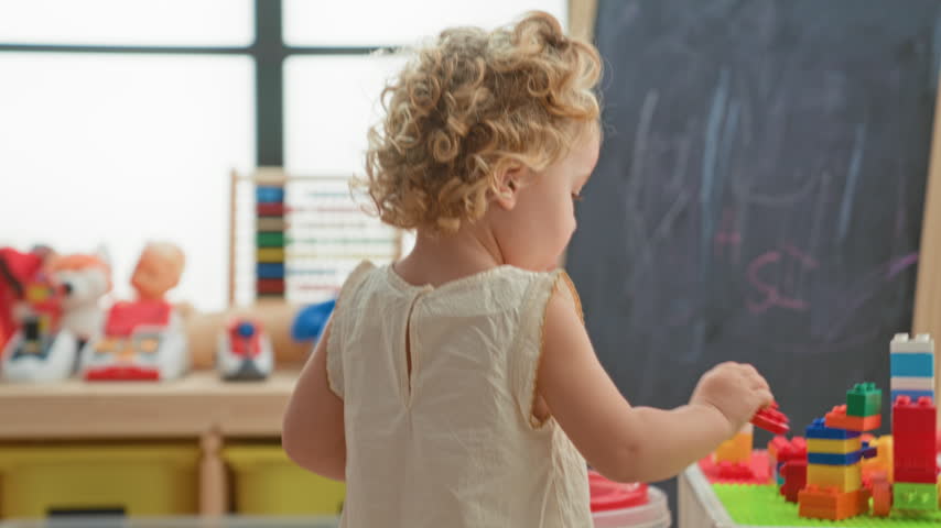 Little blonde girl holds a red lid with hand while reaching toward stacked blocks in a playroom building; curiosity.