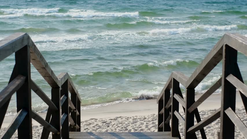 Stairs lead down to sandy beach from wooden deck with gentle waves rolling in under bright blue sky. Wooden deck frames breathtaking view of beach and water meeting horizon.