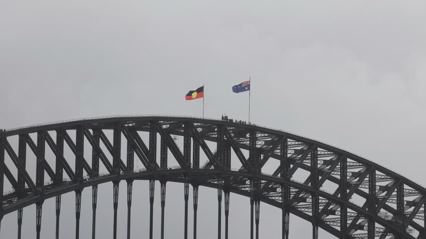 View of Sydney Harbour Bridge with the Australian flag and the Aboriginal flag flying on the steel arch under an overcast sky in Sydney, New South Wales, Australia.