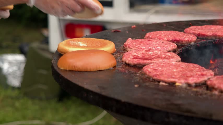 Cook toasts hamburger buns alongside raw beef patties on circular iron hotplate. Flaming wood fire heats large barbecue during sunny weekend outdoor fair