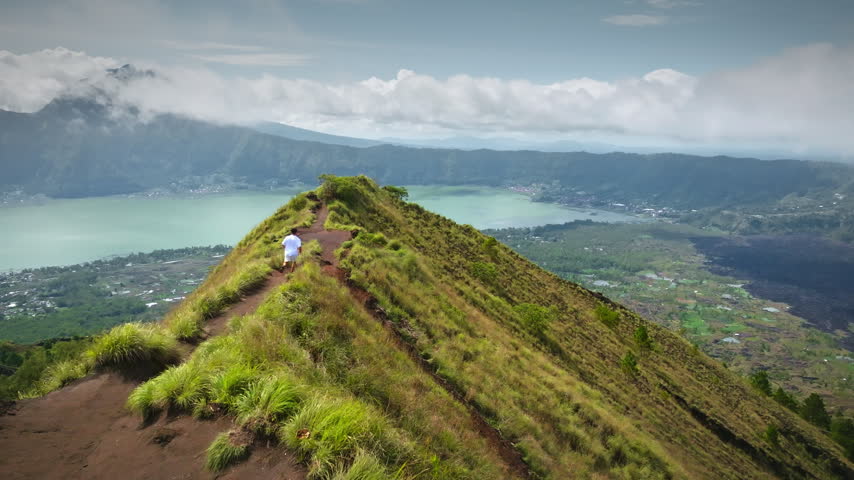 Man running along a grassy mountain ridge trail on Mount Batur, with Lake Batur and the surrounding volcanic caldera landscape visible under a cloudy sky in Bali, Indonesia. Aerial view drone flight