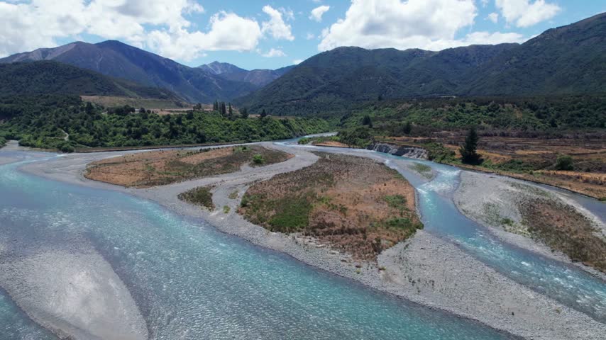 River flows through scenic landscape surrounded by mountains and forests, showcasing vibrant natural colors and diverse terrain under soft clouds in bright daylight.