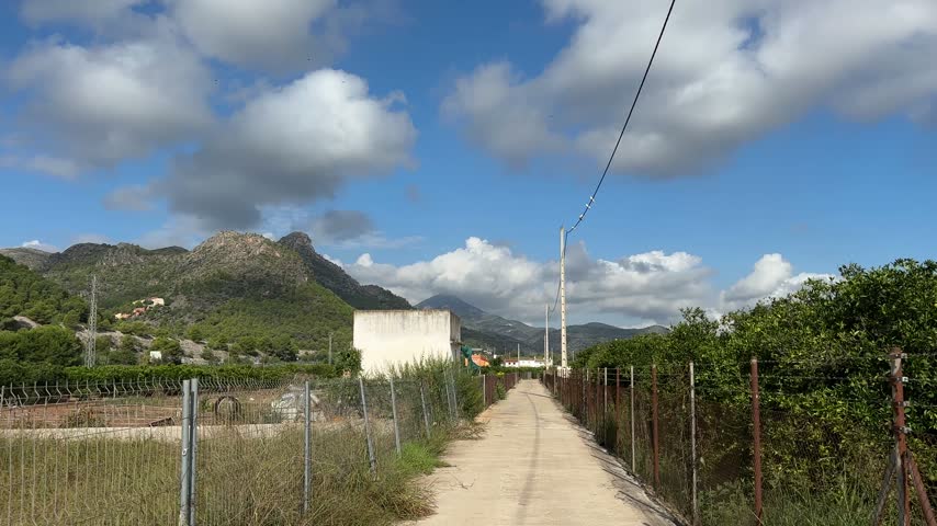 A dirt track runs straight between rows of orange trees towards hazy mountains under a sky of white clouds in Gandia, Spain.