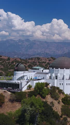 Aerial view of Griffith Observatory set against rolling hills, distant mountains, and clouds under a bright sky in Los Angeles, California, with visitors visible on the observation terraces.