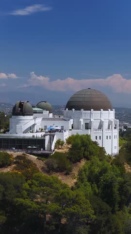 Aerial view of Griffith Observatory perched on Mount Hollywood in Los Angeles, California, with visitors walking on terraces surrounded by hillside vegetation under a bright sky.