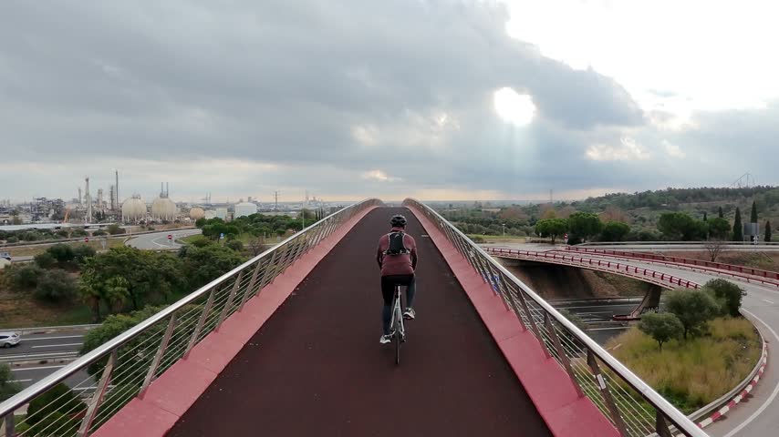 Cyclist riding along an elevated bicycle path overlooking roads, green Mediterranean landscape and an industrial complex under dramatic storm clouds with sunlight breaking through in Vila-seca, Spain.