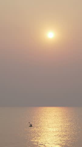 A solitary kayaker paddles across calm waters at sunrise. Misty veil hides the sun, creating golden reflections and a serene silhouette against the peaceful ocean fog.