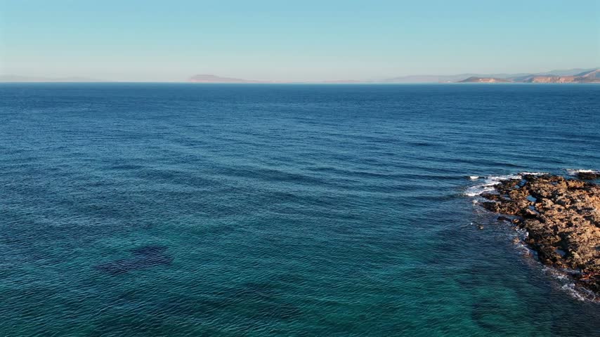 Aerial drone shot over the rocky coastline of Amorgos with the calm blue Aegean Sea and a distant hazy island on the horizon, Cyclades, Greece, on a sunny day