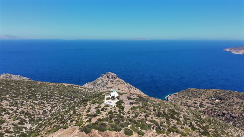 Aerial drone shot of a small blue-domed white chapel on a rocky headland overlooking the deep blue sea on Amorgos, Cyclades, Greece, on a sunny day