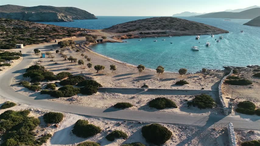 Drone view of the sandy beach and clear water of Kalotaritissa with boats and a small islet on Amorgos, Greece