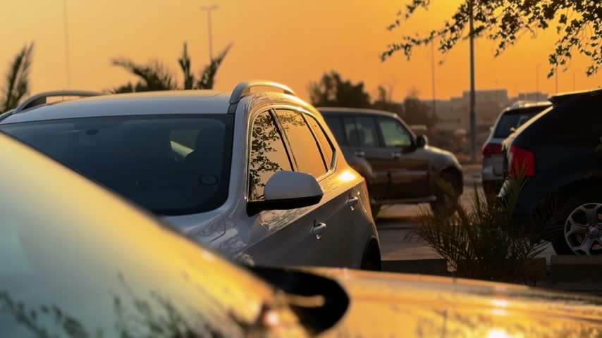 A cinematic view of parked vehicles in an outdoor lot during a vibrant golden sunset. The central silver SUV reflects silhouettes of nearby trees against the warm evening sky.