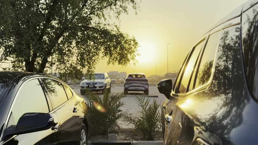 Several cars and SUVs sit parked in an outdoor lot during a vibrant golden sunset. The warm evening sunlight reflects off the vehicle windows and glossy exteriors next to green trees.