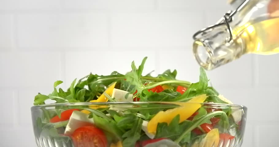 Sunflower oil pouring from bottle onto fresh vegetable salad with arugula tomato pepper and white cheese