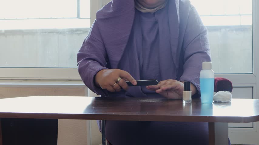 A woman is doing her manicure and filing her nails at a table in a room. Medium shot.