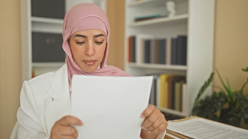 Woman in pink hijab smiling reads paper, holds document and pumps fist in office building with stacked files on desk; joy achievement.