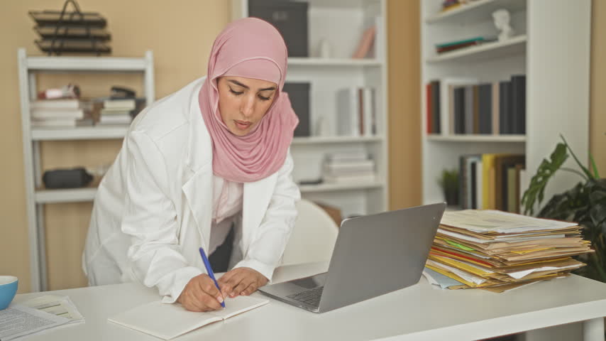 Woman in white coat and pink hijab leaning over laptop writing notes in a cluttered office with stacked files and books in building; professional focus.