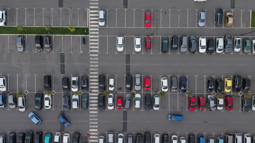 Many cars are parked in a lot beside a shopping center. People are coming and going as they shop.