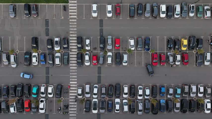 Cars are parked in a large lot. Different colors and sizes are visible. The area has marked spaces and some plants.