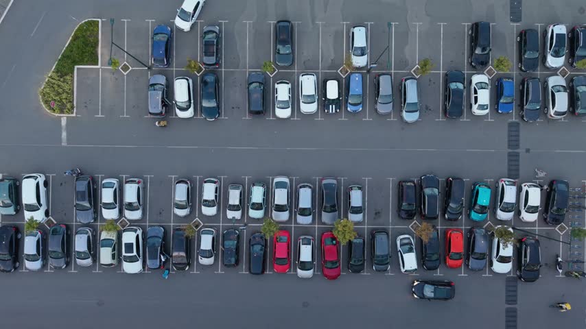 Aerial view shows many cars parked in lines in a large parking lot. The time is daytime.