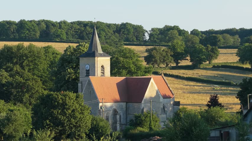 Wide shot of a traditional stone church with a clock tower and red-tiled roof in the village of Cuncy les Varzy, France. The church is nestled among lush green trees and set against rolling fields and