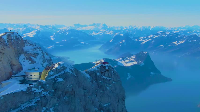 A wide aerial shot looking over a dramatic snowy mountain peak down toward a large blue lake surrounded by mountain ranges.
