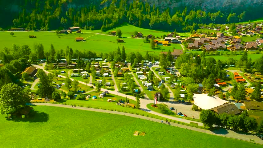 A high-angle aerial drone shot of a lush green campground filled with RVs and tents next to a charming alpine village.
