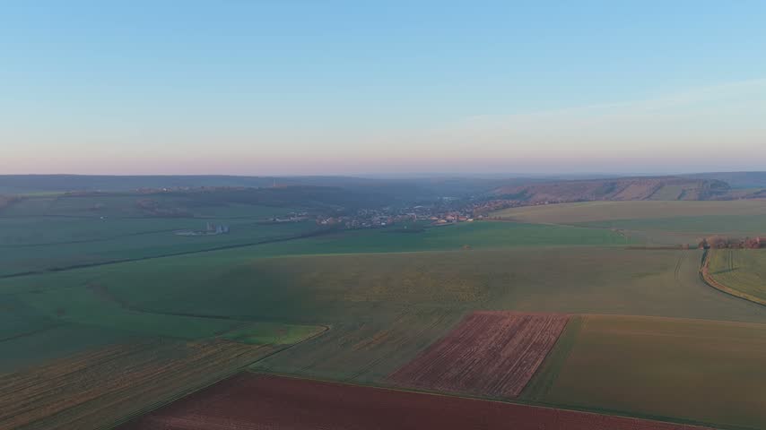 Drone footage capturing expansive agricultural fields and a small village in the rolling countryside of Nievre, France. The landscape is bathed in soft early morning light, with green and brown