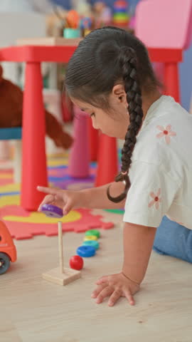 Girl kneeling and stacking colorful wooden rings with right hand on floor in building playroom among toys; concentration learning.
